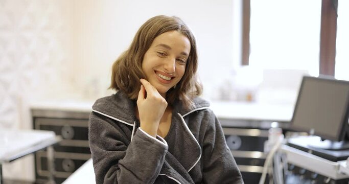Portrait of a young woman in bathrobe happy with rejuvenation procedures sitting in a beauty salon. Ultrasound lifting procedure for a face. Anti-age and wellness concept