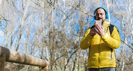 young hipster man with a beard and backpack on his back listening to music with his headphones and his mobile phone in a park