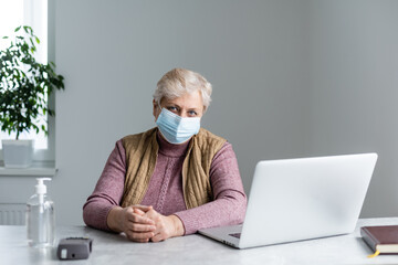 Technology, old age and people concept - lonely sad older senior woman in face medical mask working and making a video call with laptop computer at home during coronavirus COVID19 pandemic. Stay home
