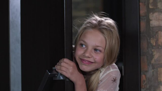 Close-up of smiling curious child opening door and enthusiastically prying on something happening in room. Cute little girl with admiration watching through crack at action taking place outside door