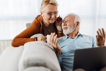 Obraz premium Cheerful senior couple relaxing on sofa in the living room and using laptop
