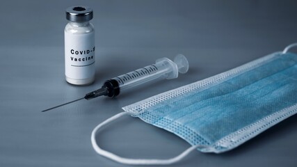 Vaccine ampoule with COVID-19 inscription, injection syringe and medical protective mask on a gray...