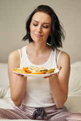 young woman with shoulder-length dark hair in light-colored pajamas looks at a plate of chopped fruits and frowns