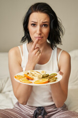 a young woman with shoulder-length dark hair in light-colored pajamas looks at a plate of sliced fruits and frowns, pressing her finger to her lips.