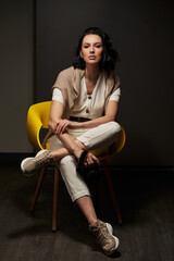 beautiful young woman in light clothes sits on a cool yellow chair in a dark interior with one leg over the other