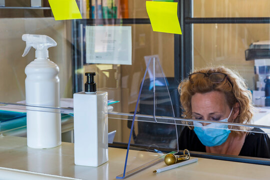 A woman with a protective mask works in an office at the reception of a hotel separated by a sheet of plexiglass