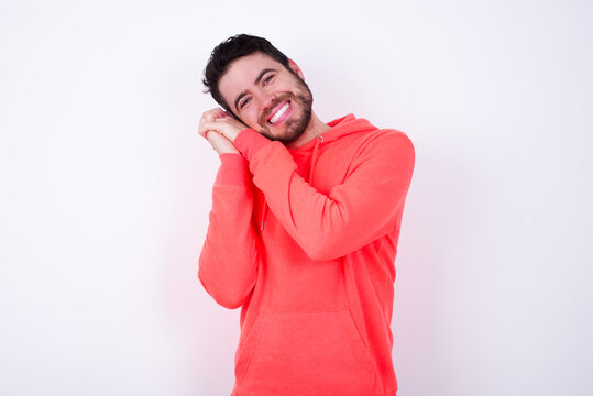 Dreamy young Caucasian bearded man wearing pink hoodie against white background with pleasant expression, closes eyes, keeps hands crossed near face, thinks about something pleasant