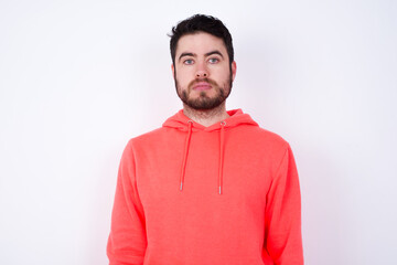 Fototapeta premium Joyful young Caucasian bearded man wearing pink hoodie against white background looking to the camera, thinking about something. Both arms down, neutral facial expression.