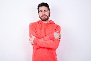 young Caucasian bearded man wearing pink hoodie against white wall bitting his mouth and looking worried and scared crossing arms, worry and doubt.