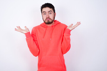 Puzzled and clueless young Caucasian bearded man wearing pink hoodie against white wall with arms out, shrugging shoulders, saying: who cares, so what, I don't know. Negative human emotions.