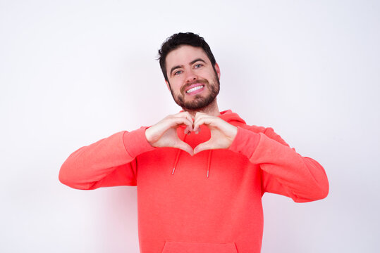 young Caucasian bearded man wearing pink hoodie against white wall smiling in love showing heart symbol and shape with hands. Romantic concept. - Powered by Adobe