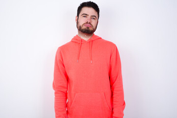 Gloomy, bored young Caucasian bearded man wearing pink hoodie against white wall frowns face looking up, being upset with so much talking hands down, feels tired and wants to leave.