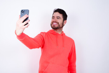 Portrait of a young Caucasian bearded man wearing pink hoodie against white background taking a selfie to send it to friends and followers or post it on his social media.