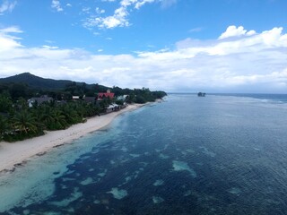 aerial view of Seychelles La Digue Island coastline with white sandy beach, green trees, blue turquoise clear water, reef and blue sky with white clouds