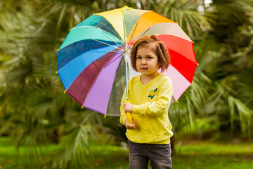 beautiful little girl.in a yellow sweater, jeans,and yellow boots, he stands with an umbrella against the background of green palm trees