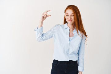 Disappointed redhead girl in blue shirt showing something small and grimacing awkward, judging something tiny or little, standing over white background