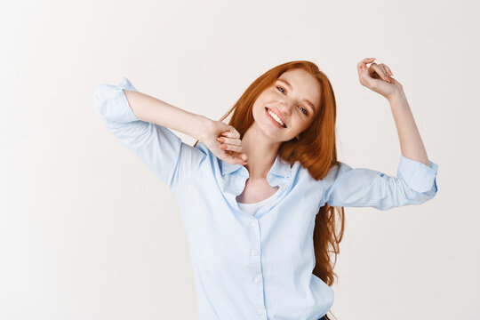 Image of beautiful girl with long red hair snap fingers and dancing, enjoying listening music, standing against white background