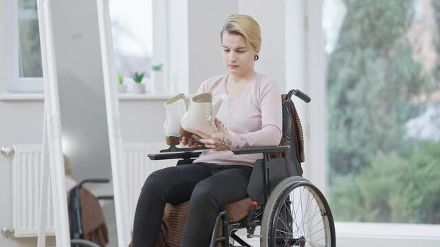 Young frustrated Caucasian woman in wheelchair looking at ice skates and thinking. Portrait of crippled paralyzed sportswoman with injured legs. Disability and paralysis.