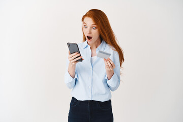 Stylish redhead girl staring surprised at smartphone screen while shopping online, holding plastic card, reaction on internet special deal promotion