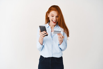 Fototapeta premium Cheerful girl shopping online, making purchase in smartphone app with plastic credit card, standing against white background