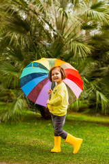 beautiful little girl.in a yellow sweater, jeans,and yellow boots, he stands with an umbrella against the background of green palm trees