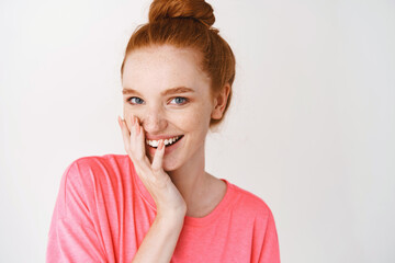 Headshot of redhead girl with messy bun, laughing and touching perfect hydrated skin, applying facial treatment and smiling, white background