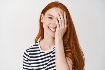 Pretty female moel with ginger hair and blue eyes smiling, covering half of face with hand, standing over white background