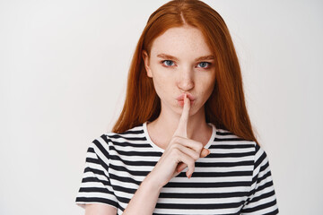 Close-up of young ginger girl with serious face showing taboo sign, hushing at you, telling a...