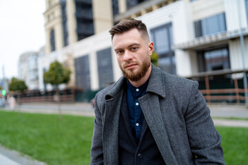 Naklejka premium Handsome and cheerful young businessman dressed in formal clothes. Male in coat is looking to the camera with city view background.