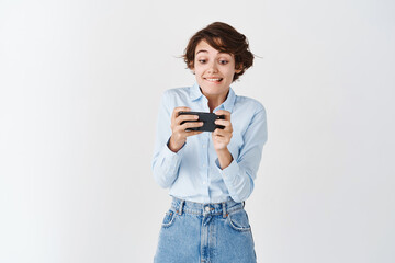 Excited female student playing video games on mobile phone and smiling at smartphone screen, standing on white background