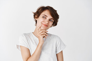 Portrait of natural young woman with short hairstyle, looking pensive aside, smiling and dreaming of something, standing thoughtful on white background
