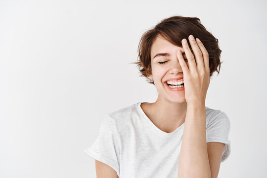 Happy european girl with short hair, cover half of face and smiling, laughing carefree, standing in t-shirt on white background