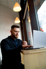 Bearded fashion young manager works on laptop while having coffee break. Modern interior of cafe. Closeup.