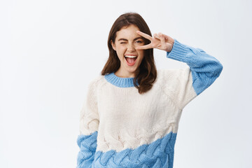 Positive young woman showing v-sign near eye and winking at camera, showing joy and happiness, feeling upbeat while standing in sweater against white background