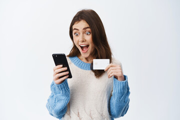 Online shopping. Excited woman staring at smartphone and holding plastic credit card with amazed face, reading sale promo offer on screen, white background