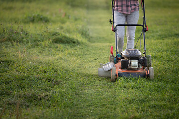 A woman in casual clothes with a gasoline lawnmower on wheels mows the grass on the lawn near the house. The powerful mower mows flowers and other plants. Improvement of the territory.