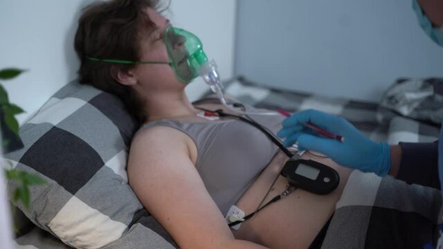Male medical worker in blue uniform and woman patient with an oxygen mask lie on bed at home. Doctor makes medical exam to quarantined female with respiratory and cardiac diseases during coronavirus