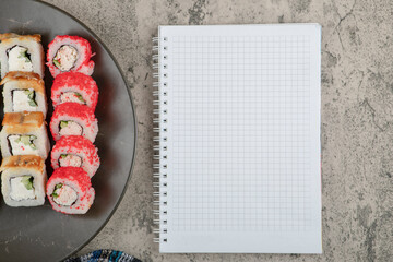 Plate of various delicious sushi rolls with empty notebook on marble table