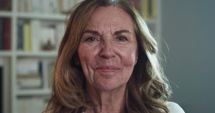 Cinematic shot of attractive happy mature senior woman is smiling in camera in a living room at home. Concept of life, family, love, mother, grandmother, femininity, women's day, mothers day.
