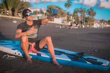 a young man uses his mobile phone sitting on his paddle board on the beach and takes a photo