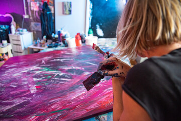 Young woman paints an abstract picture with her hands in her interior studio, focus on hand and brush