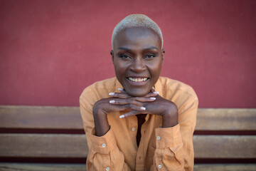 A black adult woman is sitting on a wooden bench and a red wall is in the background. She is relaxed. Focus on the face