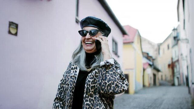 Senior chic woman with sunglasses standing outdoors, using smartphone.