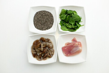 On a white plate, four plates of raw chicken, pickled mushrooms, fresh spinach leaves and chia seeds. These are plant and animal protein options.