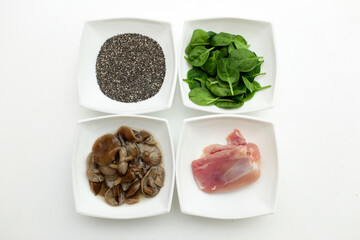 On a white plate, four plates of raw chicken, pickled mushrooms, fresh spinach leaves and chia seeds. These are plant and animal protein options.