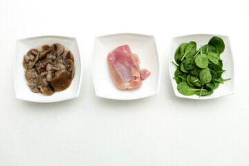 Isolated on a white background there are 3 white plates with different types of animal and vegetable. Plates include fresh green spinach leaves, chicken and pickled mushrooms. Have place for text