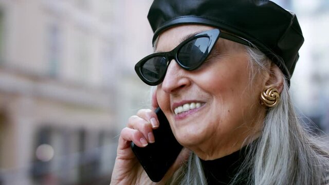 Senior chic woman with sunglasses standing outdoors, using smartphone.