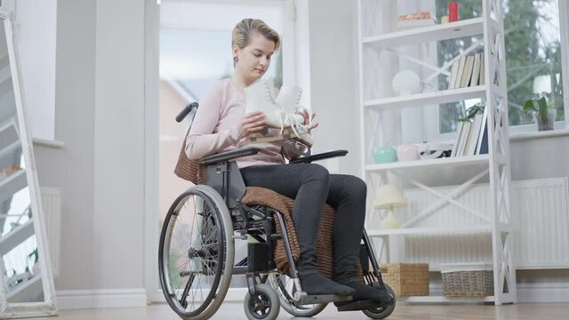 Wide shot of young depressed sportswoman sitting on wheelchair hugging ice skates. Portrait of frustrated handicapped paralyzed Caucasian woman with sports equipment at home. Disability and lifestyle.