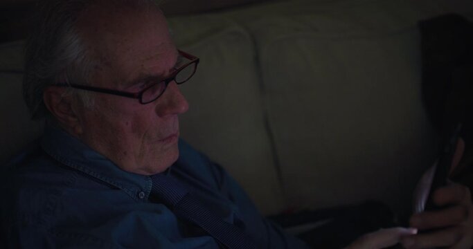 Cinematic shot of modern senior pensioner man using smartphone for entertainment while sitting on a sofa in living room at home at night .Concept: technology, modern generation, connection, retirement