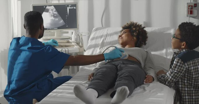 Little african kid standing near mother having ultrasound at doctor